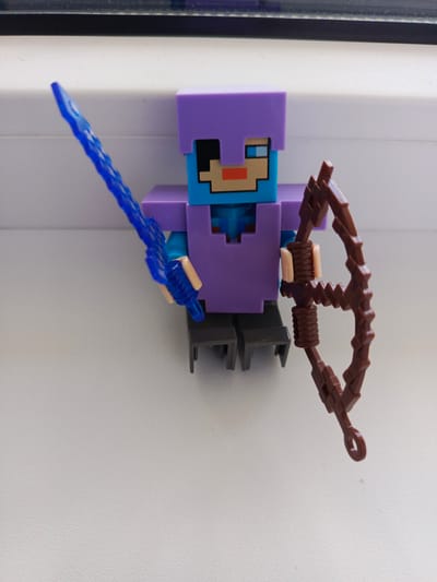 A Minecraft-themed action figure, featuring a purple helmet and armor, is positioned on a white, textured surface, possibly a windowsill or ledge. The figure holds a translucent blue sword in its left hand and a brown bow in its right. The background consists of a dark stripe, likely a window frame, above the white surface. The scene is captured from a slightly elevated angle. The location of Biaroza, Belarus, is not discernible from the visual information provided in the image.