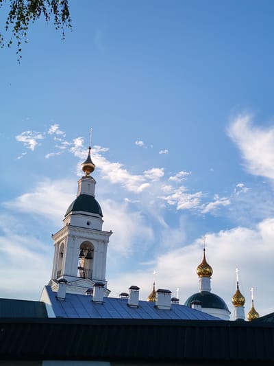 The image shows a view of a church in Vasil'ki, Russia. In the foreground, a dark-colored roofline runs horizontally across the bottom of the frame. Above this, a light blue roof covers the main structure of the building, punctuated by several white chimneys with dark caps. To the left, a tall, white bell tower rises, featuring arched openings revealing bells within, and topped with a green dome and a golden spire with a cross. Further to the right, multiple golden domes with crosses are visible, some with green bases. The sky is a bright blue, with scattered white clouds, some wispy and others more defined. A few green leaves and branches hang from the top left corner of the image. No people or animals are visible.