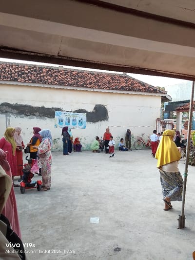 An outdoor scene captured in Jagapura Kulon, Indonesia, depicts a group of approximately 15-20 individuals gathered on a concrete surface. The setting features a cream-colored building with a distinctive reddish-brown tiled roof in the background, spanning horizontally across the upper middle of the frame. The wall of this building exhibits unevenly patched sections of darker material. A large, light-colored banner or poster featuring multiple figures and text is affixed to the center of the wall.
Individuals, predominantly adults and several children, are dispersed across the concrete area. Many women are wearing hijabs or headscarves of various colors, including yellow, green, pink, and patterned fabrics. On the left side, three women in pink and yellow attire stand near a red child's stroller or wagon. Towards the center, a woman in a light-colored floral print dress stands, while a young child in a white shirt and red shorts walks or runs towards the right. Several people are seated or crouching along the wall, including a woman in a green top and another in a red top. A red bicycle is parked near the right side of the wall. On the far right, partially obscured by a metal pole, a person wearing a bright yellow hijab and a patterned skirt stands with their back to the viewer. The concrete ground appears light gray with some visible marks and variations in texture. An elevated concrete structure or overhang partially obscures the top of the image frame. The image metadata indicates it was captured on "vivo y19s" by "Akthar & Arvan" on "15 Jul 2025, 07:26".