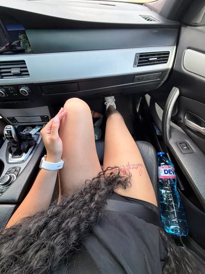 This media file captures an indoor scene within a car, likely during daylight hours as indicated by the lighting. The primary subjects are a person, partially visible, and various car interior elements. The person is seated, with their legs extended and their arm resting on the center console. They are wearing a black outfit and a white smartwatch. A noticeable detail is a tattoo on their thigh with visible red lettering.
The car interior is detailed with a focus on the dashboard and center console. Visible objects include a blue plastic water bottle with a label that says "DEVI" and some Cyrillic text, a car air vent, and the gear shift. The overall setting is functional, typical of a vehicle's interior.
The activity appears to be a moment of rest or waiting within the car. The image doesn't convey a strong sense of emotion, but the composition suggests a casual, everyday moment. The location cue of "Брусовeте, Bulgaria" provides geographical context for the setting. The lighting suggests it is daytime, but no specific weather conditions can be determined.