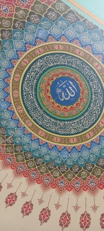 The image displays a multi-colored, intricate Islamic calligraphic artwork mounted on a ceiling or dome in the city of Kumbang, Indonesia. The artwork is dominated by a large circular design composed of concentric rings. The innermost circle, rendered in blue, prominently features the word 'Allah' (الله) in large white Arabic script. This central medallion is bordered by a ring of red and orange patterned motifs, which in turn is surrounded by a broader teal-green band inscribed with white Arabic calligraphy. Outward from these central rings, a sequence of interlocking, scalloped patterns in various shades of blue, green, and red-pink extend, each segment containing smaller white Arabic script. The lower section of the image shows a cream-colored background decorated with stylized red and pink floral drop patterns. A thin, silver-colored chain descends diagonally from the artwork's center. A light blue ceiling edge and a wooden beam are visible at the upper right, forming part of the ceiling structure. No people, actions, or interactions are present.