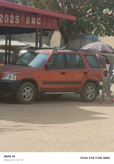 An orange first-generation Honda CR-V SUV is situated on a dirt ground in Tudun Wazurchi, Nigeria. The vehicle, equipped with a roof rack and side steps, is oriented with its front towards the left of the frame. A young male child, wearing a light-colored long-sleeved tunic, a cap, and pink sandals, walks from left to right on the dirt ground, partially obscured by the SUV.

Above the SUV, a red metal structure with "2025 KMC" painted in white lettering is visible, suggesting a permanent fixture or shelter. To the right, a large, open, dark-colored umbrella provides shade. Behind the SUV and umbrella, a partial view of a three-wheeled vehicle (likely an auto-rickshaw or tuk-tuk) and a person in a red shirt is discernible. Further in the background, a large tree with visible foliage and multiple light-colored buildings with windows are present. The scene is brightly lit by sunlight, casting shadows on the ground.