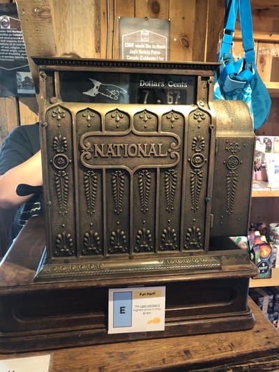 The image shows an antique National cash register displayed on a wooden counter. The cash register is made of brass with intricate decorative carvings. A sign attached to the counter in front of the cash register reads "Fun Fact!" and indicates that the cash register's highest price is only $7.99. In the background, there are wooden walls, shelves with merchandise, and a blue bag hanging on the wall. A person's arm and shoulder are visible on the left side of the image, near the cash register. The signage in the background, "CMHF would like to thank Jag's Variety Petro-Canada (Coldwater) for", suggests the location is Coldwater, which is in the city of Severn, Ontario, Canada.