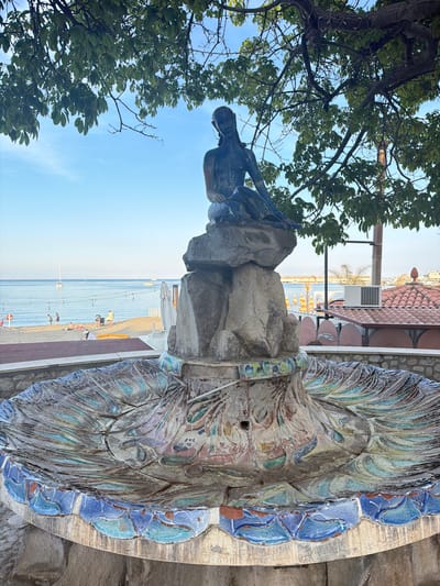The photograph, taken in Chianchitta, Italy, shows a statue atop a stone structure, possibly a fountain, with a coastline in the background. The statue is of a person, perhaps male, sitting and gazing into the distance. The structure has a decorative, shell-like design with blue and grey-toned tiles around the edge. A tree is visible to the left, partially obscuring the sky. The background reveals a beach with people, a building with a red-tiled roof, and the sea. The scene is bathed in daylight.