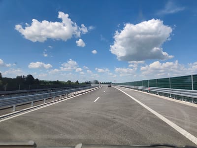 This media file captures a view of a modern highway on a sunny day, likely in or near Peshterna, Bulgaria. The perspective is from inside a vehicle, looking forward down an empty asphalt road with clear white lane markings. The highway is flanked by guardrails on both sides, with a tall, green sound barrier visible on the right. In the distance, a single dark-colored car is driving away from the camera.
The sky is a vibrant blue, dotted with fluffy white cumulus clouds, suggesting fair weather. The surrounding landscape is green and hilly, with a line of trees visible in the distance beyond the left-hand guardrail. The sun is high in the sky, indicating it is likely midday. There are no people visible in the scene, only the car on the highway. The overall impression is one of open road and smooth travel under good weather conditions. No visible text is present in the image.