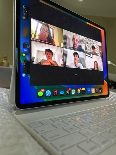 The image shows a laptop displaying a Zoom meeting. The meeting has six participants: Anabelle Helene Elizabeth Marie Herion, Baptiste Delmas, Denis Lamet, Alvaro Navarro De Andres, Carlos, and a woman whose name is not visible. The laptop is connected to a keyboard. The scene takes place in Río Oro, Costa Rica at the coordinates 9.945, -84.201. The image is taken from a low angle and shows the laptop in focus, while the background is blurred. The laptop screen is displaying a Zoom meeting, with the participants' names and video feeds visible. The keyboard is white and has a standard layout. The laptop is on a white surface and there is a white object in the background.