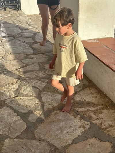 A young child, approximately three to five years old, is depicted in the foreground, walking barefoot on a sunlit, irregularly stone-paved path. The path consists of light-colored flagstones set with visible sand and gravel between them. The child wears a loose-fitting, light-colored (possibly cream or light yellow) t-shirt with red text reading "COPACABANA Rio Janeiro FROM SUNRISE SAMBA" and striped shorts. The child's head is angled downwards and to the left, with brown hair partially obscuring their face. Rosy cheeks are visible. In the background, behind the child, the lower legs and bare feet of an adult, wearing dark shorts, are partially visible. To the right of the child, a light-colored building wall rises, with a terracotta-tiled ledge or step running along its base. Distinct shadows are cast on the ground, indicating bright sunlight. A portion of a black wheeled object, possibly a stroller, is visible in the upper left corner. The scene takes place outdoors in Palafrugell, Spain.