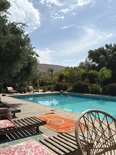 This image depicts a serene outdoor swimming pool area, presented on its side, but intended to be viewed in portrait orientation. When correctly oriented, the scene is dominated by a bright, partly cloudy sky with scattered white cirrus clouds at the top.
The right side of the frame features a clear, light blue swimming pool, reflecting the sky and surrounding foliage. Adjacent to the pool is a paved area where several wooden lounge chairs with colorful, floral-patterned cushions are arranged. A vibrant, ornate rug, primarily in shades of red and orange with intricate patterns, is laid out on the ground near the seating. Strong, angular shadows are cast by the chairs and other poolside furniture, indicating bright sunshine, likely late morning or early afternoon. A large, circular, woven chair back is partially visible in the bottom right corner.
Beyond the pool and its immediate surround, a dense expanse of lush green trees and shrubs forms a natural border, creating a vibrant midground. These include both broadleaf trees and palm-like plants. In the far background, rising majestically above the greenery, is a large, arid mountain range, characteristic of the landscape around Tahannaout, Morocco, under the expansive blue sky. No people or visible text are present in the image. The overall scene evokes a peaceful and relaxing ambiance.