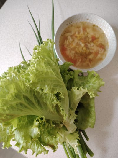 A bunch of fresh green lettuce and green onions is placed in the foreground on a speckled white surface. In the background, a white bowl with a light blue geometric pattern contains a clear broth soup with visible vegetables and herbs. The scene is well-lit, suggesting it might be indoors, possibly in a kitchen or dining area, in a location such as Кривец, Russia, though the specific location cannot be confirmed from the image alone.