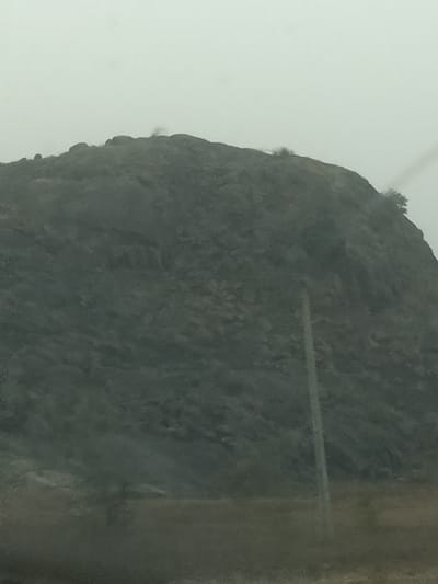 The scene, located in Barakin Tenti, Nigeria, features a large, dark, rocky mountain dominating the majority of the frame under an overcast, light gray sky. The mountain exhibits rugged terrain with visible rock formations and sparse, dark green vegetation, including small trees and bushes, primarily along its crest and upper slopes. A single, slender, light-colored pole, consistent with a utility pole, extends vertically from the lower frame upwards, positioned in the mid-right foreground against the mountain's base. The immediate foreground presents an indistinct, dark ground area. No persons, animals, or specific activities are observed.