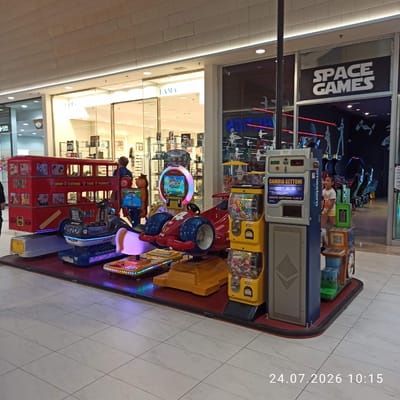 The image shows a row of colorful children's amusement rides situated in a brightly lit indoor shopping mall, likely a game arcade. In the foreground, a bright red double-decker bus ride is prominent, adorned with various cartoon images and destination names such as "LONDON", "OXFORD", "YORK", and "WEMBLEY". Adjacent to it are other rides, including a red race car with blue wheels and a purple-lit platform. A yellow vending machine with "PAWS" written on it is also visible. In the background, a section of a store with the text "SPACE GAMES" is visible through a glass window, suggesting the amusement rides are part of or adjacent to this gaming establishment. Another store with the signage "LAMA" is also visible. The flooring consists of light-colored, square tiles. A timestamp "24.07.2026 10:15" is overlaid in the bottom right corner of the image. The location is identified as Modugno, Italy.