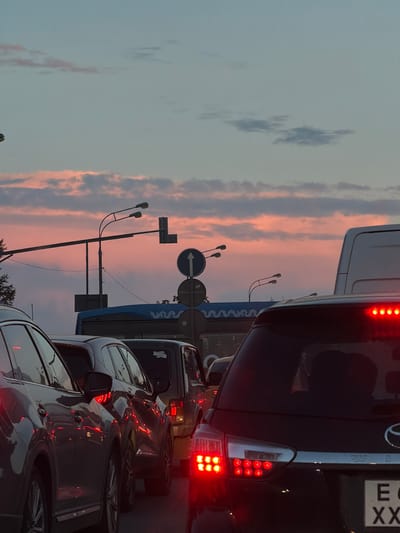 This is a vertical, medium-shot, outdoor photograph taken at dusk in the city of Krasnaya Pakhra, Russia. The image depicts a traffic jam on a multi-lane road. In the foreground, the rear of a black SUV is visible, with its brake lights illuminated. To the left, the side of a grey SUV is seen. Behind these vehicles, other cars and a bus are stopped in traffic. Above the vehicles, a street light with two lamps is mounted on a pole. Traffic signs, including a circular sign with a white arrow pointing upwards and a rectangular sign, are also present. The sky is a gradient of pale blue and pink, with scattered clouds, indicating the sunset. The overall scene conveys a sense of urban congestion during evening hours.
