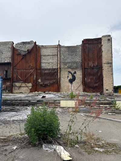 A partially demolished brick building with a mural painted on the side depicting a woman in a black dress doing a ballet pose. The building is located at 50.561° N, 30.31° E in Horenka, Ukraine. The building is made of brick and has a large, rusted metal sheet covering a portion of the wall. The wall is damaged and is missing some sections of brick. The mural is painted in black and white and is in good condition. There is a section of rubble and broken concrete between the building and the foreground of the image. The foreground is dominated by a small bush and a few scraggly weeds growing in the dirt. The sky is overcast with a hazy appearance.