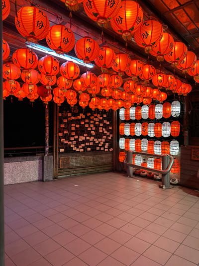 The image captures an illuminated covered walkway or plaza at night, likely in Jiufen Old Street, New Taipei City, Taiwan. The overhead structure is densely covered with hundreds of glowing red paper lanterns, predominantly round with black Chinese characters, suspended in rows. These lanterns are the primary light source, casting a warm, orange hue over the scene.
On the far right, a section features multiple vertical rows of cylindrical lanterns. These alternate between white and orange, displaying text such as "TAIPEI" and other phrases in various languages. Below these, a silver-colored metal barrier, consisting of two vertical posts supporting a curved horizontal rail, stands on the tiled floor. Beneath this barrier, several spherical, dark-colored objects, possibly decorative lanterns or gourds, are arranged on the ground.
To the left, a low, speckled grey concrete barrier or wall separates the walkway from an unlit, dark exterior. Adjacent to the right, a dark-toned wall is covered with numerous small, light-colored rectangular papers, possibly wish slips or notes, attached in a grid pattern. The floor is paved with light-colored square tiles. A single, crumpled paper cup is visible on the tiled floor near the base of the metal barrier. No individuals are present in the scene.