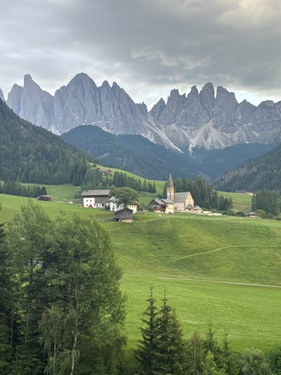 This is a vertical, eye-level shot of a picturesque mountain village in St. Magdalena, Italy. The scene is dominated by dramatic, jagged gray mountains rising sharply in the background under a cloudy sky. In the middle ground, a cluster of rustic buildings, including a church with a prominent clock tower, are nestled on a lush green hillside. The village is surrounded by dense green forests and expansive, rolling meadows. A paved path winds through the meadows, suggesting a tranquil setting ideal for hiking or leisurely strolls. In the foreground, a thick canopy of green trees frames the view. The weather appears overcast, creating a soft, diffused light that highlights the vibrant greens of the landscape. No people are visible, and no text is present in the image. The overall mood is one of serene natural beauty and peaceful village life.