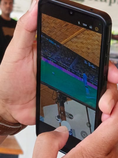 In Julikeude Dua, Indonesia, a close-up shot captures a person's hands holding a smartphone. The phone's screen displays a video game simulation of a soccer match, complete with players on a green field and stadium seating. The top status bar of the phone shows a lightning bolt icon indicating airplane mode, the number "12M," a clock icon, and a smiley face emoji. The camera interface is also visible on the screen, with zoom options like "0.5" and "2.8x" displayed. The background is blurred, but a person in a dark shirt can be partially seen. The scene is brightly lit, suggesting an outdoor or well-lit indoor environment.