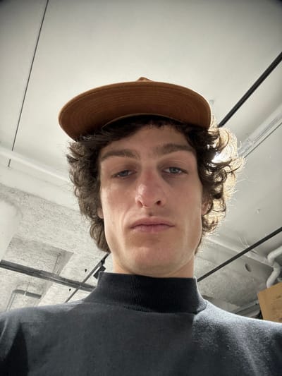 A male individual is depicted in a low-angle, close-up photograph taken indoors in Joinville-le-Pont, France. The subject, appearing to be an adult male, has curly dark brown hair and light-colored eyes, with a small metallic ring piercing on the right nostril. He wears a black mock neck shirt and a flat-brimmed, brown-toned hat resting atop his head. His expression is neutral, and he is looking directly towards the camera. The background reveals an interior space with a light-colored, textured ceiling, featuring dark linear structural elements or tracks, and white pipes. Illumination appears to be overhead, creating a backlight effect on the subject's right side.