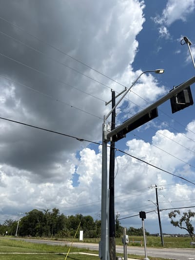 The image depicts an outdoor scene in Parrish, United States, dominated by a cloudy sky with patches of blue. In the foreground and mid-ground, several utility poles and power lines are visible, stretching across the frame. One pole, a prominent vertical element, supports traffic signals and a street light. The traffic signals are black and rectangular, and the street light is illuminated. Another utility pole further back also carries power lines and a street light. The ground in the foreground is covered with green grass, and a road is visible in the mid-ground. Beyond the road, there is a line of dense trees. In the background, residential structures and a body of water can be seen. The overall impression is that of a street scene on a partly cloudy day.