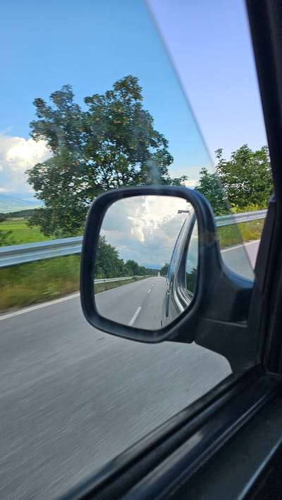This media file presents a dynamic view from the interior of a moving vehicle, looking out the side window and into the side mirror. The overall composition is tilted, giving a vertical orientation to the landscape beyond the car.

Through the upper part of the side window, a clear, bright blue sky with scattered white clouds is visible, alongside the verdant green canopy of a large tree. The lower section of the direct view shows more of the tree and a glimpse of green fields. A silver guardrail runs along the roadside.

The black-framed side mirror occupies a prominent central position, reflecting the road behind and to the immediate side of the vehicle. Within the mirror's reflection, a grey asphalt road stretches into the distance, marked by a solid white shoulder line and a dashed white line separating lanes. On both sides of the reflected road, dense green trees and foliage create a natural boundary. The sky reflected in the mirror is also bright blue with fluffy white clouds, mirroring the direct view. Parts of the car's dark interior frame and door are visible on the right side of the image, indicating the perspective is from within.

The scene captures a daytime moment, characterized by clear, sunny weather with some cloud cover, suggesting pleasant conditions for travel. The blurred appearance of the road surface, particularly in the mirror's reflection, indicates the vehicle is in motion. No individuals are visible, nor is any discernible text present in the image. The setting is a rural or semi-rural highway, with the provided location context being Kliment, Bulgaria.