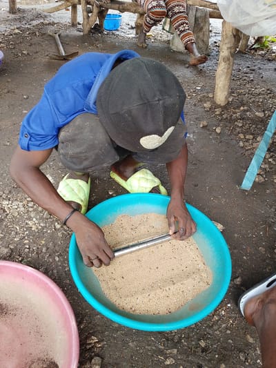 In Kajola, Nigeria, a person is crouched down, preparing to sort through a bowl of sand. The person is wearing a blue shirt, dark pants, and light green slippers. They are holding a metal object, possibly a tool, which they are using to mix the sand. The sand is in a large, bright blue bowl. Next to the blue bowl is a pink bowl, which also appears to contain sand or a similar granular substance. The ground around the bowls is dirt and gravel. In the background, there are wooden structures, possibly part of a shelter or market stall, and a blue bucket. Another person's legs, wearing patterned pants, are visible in the background. The scene suggests a process of material sorting or preparation, common in many outdoor markets or workshops in Nigeria.