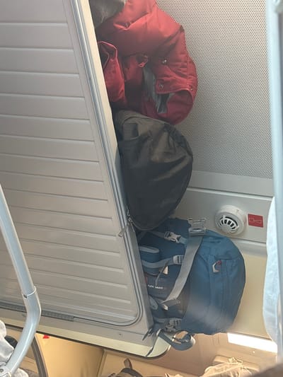 The image displays an interior view of a storage compartment, consistent with a train or transport cabin. The exact location within Druzhino, Russia, cannot be determined from the visual information provided.

The scene is dominated by a light-colored, vertically slatted compartment door, which is partially open on the left side, revealing items stored within. Inside, from top to bottom, are a crumpled red puffer-style jacket, a black soft-sided bag with a zipper pull, and a blue backpack featuring multiple gray straps, buckles, and a label indicating "FLEX VARIO."

The background wall behind the compartment consists of a light-colored perforated panel in the upper section and a smooth white panel below it. Mounted on the white panel, to the right of the compartment, is a circular white smoke detector or fire alarm with vents. Adjacent to the alarm is a red rectangular symbol, possibly a fire safety or "no smoking" indicator. Below the fire alarm, a warm-toned linear light fixture is visible, casting illumination into the lower part of the scene. A light-colored metal bar or railing is partially visible on the far left. The bottom of the image shows a blurred section of what appears to be an upholstered surface, possibly a seat or bed. No individuals are present in the image.