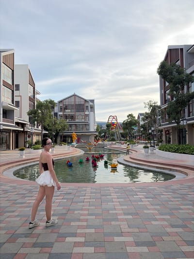 A young woman wearing a brown tube top and a white ruffled skirt stands on a patterned brick walkway in front of a large pond. She is wearing glasses and white sneakers with black stripes. In the pond, there are numerous colorful, origami-like decorations floating. The pond is surrounded by a curved walkway with planters. Behind the pond are multi-story buildings with modern architecture, featuring wood accents and large windows. Trees and a playground structure with a red arch are visible in the background. A person wearing a conical hat is walking near the pond. The sky is overcast. The scene appears to be an outdoor public space in a town or city.