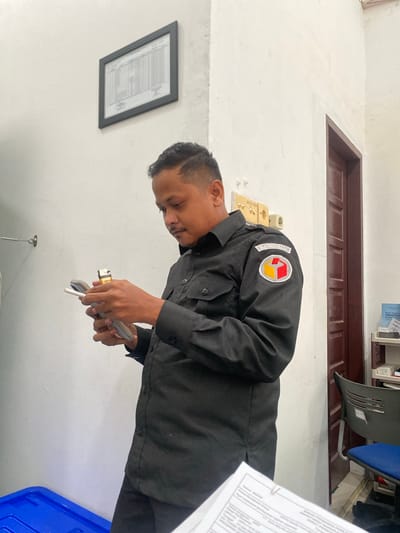 A medium shot captures a man in a black uniform, likely a security guard or official, standing indoors and focused on holding a cigarette and a lighter in his hands. The uniform features a patch on the left sleeve with the text "BAWASLU" and a circular emblem containing red, yellow, and white elements. He appears to be in the process of lighting his cigarette.
The background reveals a room with white walls. On the left wall, a framed document with a grid of text is hung. To the right, a dark wooden door is visible, and further to the right, a shelf unit holds various items, including a stack of documents and a blue chair with a grey seat. A blue plastic container is visible in the lower left of the frame. The overall impression is that of a utilitarian office or storage space. The presence of the "BAWASLU" patch suggests a connection to the Indonesian Election Supervisory Body, potentially indicating the location is an office or facility associated with this organization in Lhokseumawe, Indonesia.