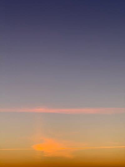 This is a vertical, full-frame photograph of a sunset sky over La Teste-de-Buch, France. The sky is a gradient of colors, transitioning from a deep indigo at the top to a warm orange at the bottom. There are faint, thin clouds present, with one prominent band of pinkish-orange clouds stretching horizontally across the middle of the frame. Several faint contrails from aircraft are visible as thin white lines against the colored sky. The overall scene is serene and devoid of any human presence or discernible man-made structures.