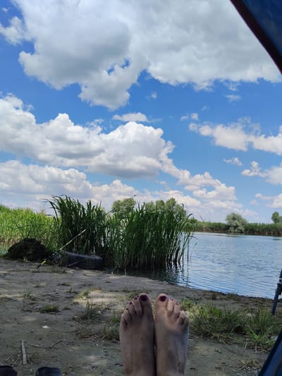 A first-person perspective image shows a person's feet in the foreground, positioned on a sandy bank next to a body of water. The feet are bare, with toenails painted a dark purple. Behind the feet, tall green reeds grow along the edge of the water. A fishing rod is visible leaning against a pile of green material on the bank, near a black tire. The water is calm, with ripples reflecting the bright blue sky. The sky is filled with fluffy white clouds. In the background, more reeds and trees line the opposite bank of the water. The scene appears to be a natural setting, possibly a lake or river. The overall mood is relaxed and tranquil, suggesting a leisurely activity like fishing on a sunny day. The location is identified as Bilyaivka, Ukraine. The time of day is likely daytime, given the bright sunlight and blue sky. The weather appears to be pleasant, with a mix of sun and clouds. No text is visible in the image.
