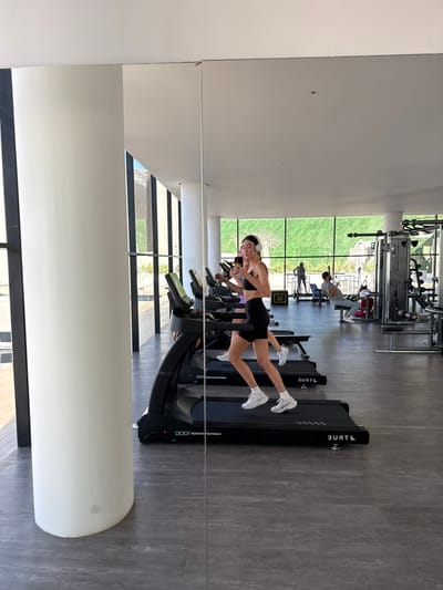 The image depicts the interior of a modern fitness center in San José del Cabo, Mexico. A long, floor-to-ceiling mirror dominates the right side of the frame, reflecting the gym's activity. In the foreground of the reflection, a young adult female with long dark hair, wearing a black sports bra, black athletic shorts, and white athletic shoes, is actively running on a black "TRUE PERFORMANCE 800" treadmill. She wears white over-ear headphones and holds a smartphone in her left hand, pointed towards the mirror, indicating she is photographing or videoing the scene.
Behind her in the reflection, another individual, partially obscured, is also running on a treadmill, wearing a purple top. Further back and to the right, a male individual is seated on a weight machine, engaged in exercise. Additional gym equipment, including other treadmills and various weight machines, is visible throughout the mirrored reflection. The gym floor is a dark gray, textured material.
To the left of the mirror, a large, white circular support column extends from the floor to the ceiling. Beyond this column, the actual space of the gym features expansive floor-to-ceiling windows with dark frames, allowing significant natural light into the facility. Through these windows, a vibrant green landscaped area is visible, suggesting an outdoor environment immediately adjacent to the building. The overall scene is brightly lit, indicative of daytime.