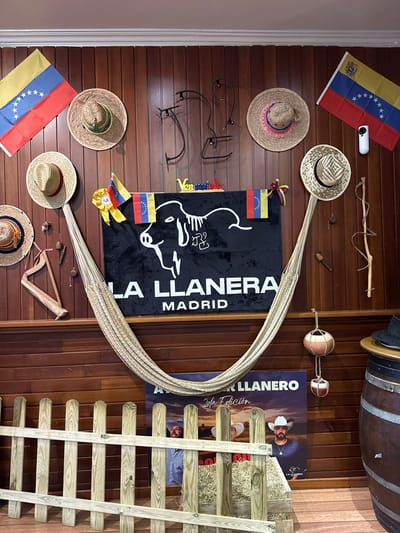 The image shows a wall decorated with Venezuelan flags, straw hats, traditional Venezuelan items like a small harp and spinning tops, and a large black banner with a white illustration of a cow and the text "LA LLANERA MADRID". A hammock is hung from the wall. In the lower part of the image, a wooden fence is in front of a poster featuring two men in cowboy hats. A barrel is visible on the right. The overall impression is a display of Venezuelan culture, possibly in a restaurant or cultural center. Torrejón de Ardoz, Spain is mentioned as the location of this establishment.
