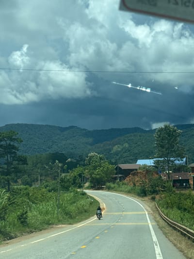 The image shows a rural road in Tu Póh, Vietnam, curving through lush green vegetation and rolling hills. The sky is dramatic, with dark, heavy clouds casting a moody light over the landscape. Sunlight breaks through in some areas, creating bright streaks. A single person rides a motorcycle on the paved road. On the right side of the road, there are some small buildings and a brown guardrail. The overall impression is one of a slightly overcast day in a serene, natural setting.