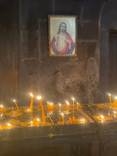 The image depicts an interior scene, likely within a religious building in Bjni, Armenia. On the left, a framed painting of Jesus Christ hangs on a textured stone wall. Jesus is shown with a halo, long dark hair, and is wearing a red robe over a white garment. He is gesturing with his right hand. The wall itself is constructed of large, rough-hewn grey stones with visible mortar lines.
To the right of the painting, a vertical metal structure is covered in numerous lit yellow candles. The candles are of varying lengths and appear to have dripped wax down the metal. The flames from the candles cast a warm, flickering light on the surrounding surfaces, creating a sense of solemnity and devotion. The density of the candles suggests they have been lit for some time. The background on the right is dark, with the illuminated candles being the primary focus. The overall atmosphere is one of quiet contemplation and religious observance. There is no visible text in the image.