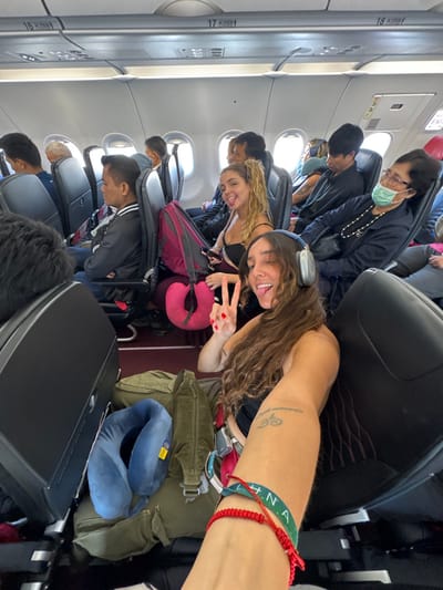 Inside an airplane, a woman with headphones poses with a peace sign. Next to her, another woman smiles. Passengers fill the seats, some wearing masks. Overhead compartments line the walls. Luggage and neck pillows are visible on the seats and floors.