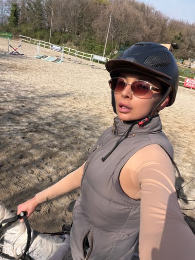 A medium, high-angle shot shows a person riding a horse in an outdoor arena. The location is Grad Labin, Croatia, based on the "Iber Pan" sign visible in the background. The rider, wearing a black riding helmet with a brown brim and round sunglasses, is clad in a light tan long-sleeved undershirt and a grey vest. They are holding the reins of the horse, which is mostly white with some grey markings. In the background is a set of horse jumping obstacles, along with trees without leaves in the far distance. The ground of the arena is sandy.