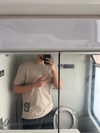A male individual is reflected in a mirror, taking a selfie inside a confined space consistent with a train lavatory, located in Stambolovo, Bulgaria. The subject, seen from the chest up, has a short beard and is wearing a plain, light beige short-sleeved t-shirt. A black smartphone with a cracked screen protector is held in his right hand, used for taking the photograph. His left hand is raised, displaying a "peace sign" with the index and middle fingers. A pink bracelet is visible on his left wrist, and a dark tattoo is partially visible on his left forearm. In the background reflection, a white toilet with an open lid is positioned on the right side. To the left, a light-colored wall features a metallic door handle and two small warning signs. On the right, beyond the toilet, a window with streaked glass shows an indistinct exterior. The overall environment features light-colored walls and overhead panels.