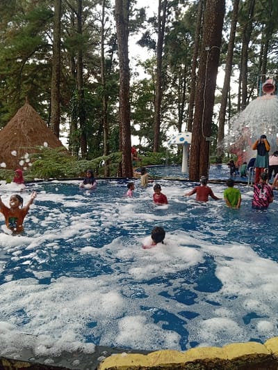 This image depicts an outdoor swimming pool facility located in Kuwukan, Indonesia. The scene is dominated by a large, blue-tiled pool densely covered with white foam. Numerous tall pine trees form the background, creating a forested environment around the pool area. Approximately ten individuals, predominantly children, are present within the foam-filled water, engaged in recreational activity. One child in the left foreground, wearing an orange shirt, has arms raised. Other individuals are partially submerged in various positions. On the right, a large cascading water feature is active, and an adult-figure wearing a light blue top stands nearby. Additional objects include a conical, thatched-roof hut on the far left and a large white decorative mushroom with blue spots positioned behind the pool in the mid-ground. The immediate foreground shows the yellow-edged border of the pool.