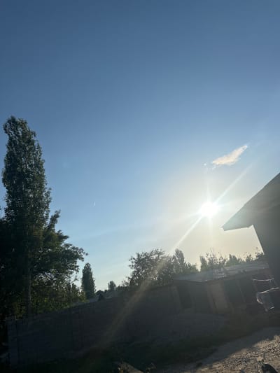 The image shows a bright, daytime sky above what appears to be a building and trees. The sun is shining intensely, causing lens flare. A small, wispy cloud is visible to the left of the sun. There is a faint contrail visible in the lower center of the sky. The building is on the right side of the image and seems to be made of dark-colored stone or concrete with multiple levels. A dark gray or black gravel area appears adjacent to the building. Trees with green foliage are visible along the bottom and right side of the image. The location is noted as Mankent, Kazakhstan.