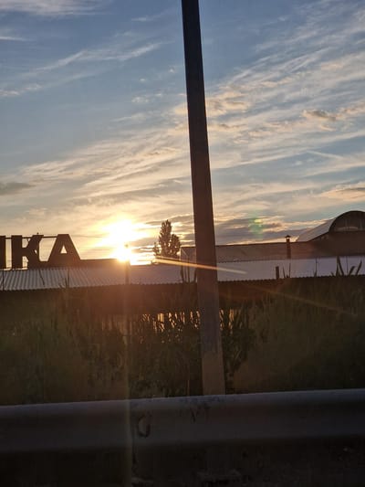The image depicts a sunset scene in Zmeyka, Russia. The sun is low in the sky, casting a warm glow through thin, wispy clouds against a blue sky. In the foreground, a metal guardrail runs horizontally across the lower portion of the frame. Behind the guardrail, dense foliage is visible. In the mid-ground, a corrugated metal roof of a building is apparent, with large, dark letters spelling out "IKA" positioned on top. To the right of the building, a tall, dark silhouette of a tree stands out against the bright sky. A vertical pole, likely a utility pole, is positioned to the left of center, obscuring a portion of the building and the sky. The overall impression is a somewhat hazy, sun-drenched rural or semi-urban landscape.
