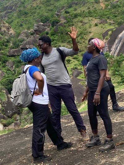Three people are standing on top of a large, sloping rock face in Ganawuri, Nigeria. The rock is gray and textured. Green vegetation covers the terrain below. The person nearest the foreground is wearing a white t-shirt, black pants with a white stripe, a blue head covering, and a gray backpack. The person in the middle is wearing a gray t-shirt and dark pants, with their left arm raised. The person furthest from the foreground is wearing a dark gray t-shirt, black leggings, and a patterned head covering. All three individuals are wearing athletic shoes. They appear to be standing near the edge of the rock, overlooking the surrounding landscape.