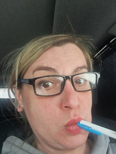 An adult female is depicted in a close-up, frontal portrait, situated within what appears to be the interior of a vehicle in Novaya Derevnya, Russia. She has blonde hair, wears black-framed eyeglasses, and a small, spherical stud earring is visible in her left earlobe. Her eyes are wide open, directed towards the camera, with visible light reflections in the pupils. Her lips are pursed and coated with reddish lipstick, actively gripping a white pen with a blue segment horizontally between them. She is dressed in a grey hooded garment, partially visible. The background is a dark, textured fabric consistent with a car's headliner, with a dark structural element visible in the upper right.