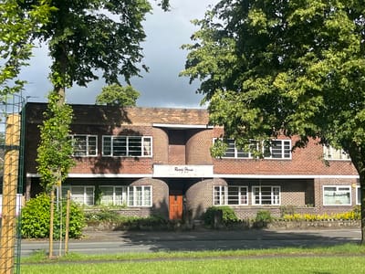 This image shows the front exterior of "Reade House", a brick building located at 12-34 Reade Street, Stretford, Trafford, United Kingdom. The building has a distinctive architectural style with curved brickwork above the entrance and rows of white-framed windows. The entrance itself features a wooden door. The building is partially obscured by lush green trees on either side. In front of the building, there is a low stone wall with yellow flowers growing along it. A paved area with a road is visible in the foreground, and beyond that, a grassy verge. The sky above is overcast with dark clouds. A metal fence and a small, young tree on a stake are visible on the far left of the image. There are no people or animals visible in the scene.