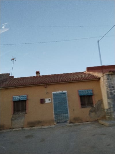 A low-angle shot showcases a single-story, tan-colored building beneath a clear, light blue sky. Red tiles cover the roof, punctuated by a small chimney. The building's façade features two windows with closed, blue blinds, a centrally located door, also with blue blinds, and a small square object on the wall between the door and the window on the left. A tall antenna stands atop the building, and a partially visible, grey stone structure adjoins on the right. The foreground consists of a grey asphalt surface.