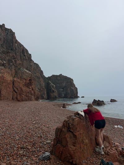 A vertical outdoor shot depicts a rocky beach scene on a grey, overcast day. A woman with blonde hair, wearing a red shirt and black shorts, is bent over a large reddish-brown rock near the water's edge, possibly looking at something in the water. The beach is composed of small reddish-brown pebbles. To the left, a large, dark, rocky cliff rises dramatically, extending into the water. The sea is calm and grey, blending into the overcast sky. Debris, including plastic bottles and a white object, are visible on the beach near the woman.
The location is Russkiy Island, Russia.