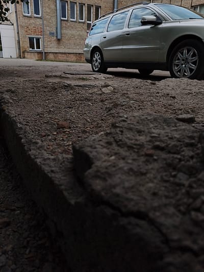 The image depicts a low-angle shot of the ground in front of a light-colored Volvo station wagon parked on a paved area next to a brick building. The foreground is dominated by rough, broken concrete and gravel, with the car and building slightly out of focus in the background. The building appears to be a commercial or institutional structure with multiple windows.

The scene is outdoors, and the lighting suggests it is daytime, possibly late afternoon or early evening, as there are shadows present. The weather appears overcast or the lighting is subdued, contributing to a somewhat somber mood. The location is identified as Tukums, Latvia. There is no visible text in the image. The primary subjects are the car, the building, and the textured ground. No people are visible, and no specific activity is taking place; the scene captures a moment of stillness. The focus on the broken ground in the foreground, contrasted with the orderly presence of the car and building, might evoke a sense of urban decay or neglect in a specific part of the location.