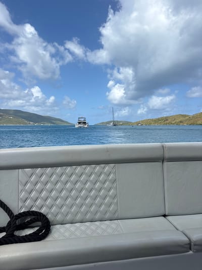 The image depicts a maritime scene within a channel or bay off Spanish Town, British Virgin Islands, under a partially cloudy, blue sky. The viewpoint is from onboard a vessel, with a light gray marine vinyl bench seat occupying the lower portion of the frame. This seat features a diamond-quilted backrest and a coiled black rope resting on its left side.
In the midground, the blue water shows slight ripples. A white, multi-deck motor yacht with a visible stern is positioned in the center-left, moving away from the viewer and leaving a wake. Further to the right, a white sailboat with a mast is present. A single red navigational buoy is visible in the water between the motor yacht and the sailboat.
The background consists of verdant, hilly landmasses on both the left and right sides of the frame, outlining the waterway. The sky above is clear blue with scattered white cumulus clouds. No individuals are clearly discernible in the scene.