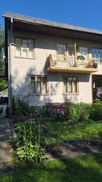 The image shows a two-story brick building with a balcony on the upper floor, located in Ļūbaste 0001, Latvia. The building has light-colored brickwork and wooden window frames with white curtains. The roof is dark and appears to be tiled. A satellite dish is attached to the exterior wall on the left side of the building.

In front of the building, there is a garden area with various plants and flowers. A wooden bench is situated in the garden, in front of a lower window. The garden is bordered by a concrete pathway on the left. Some of the plants appear to be hostas, and there are also various flowering plants in pink, white, and purple hues. The ground is covered with grass and small white flowers.

To the right of the building, a section of a car is visible under a covered area. The sky above is clear and blue. The overall scene depicts a residential property in a rural or semi-rural setting. There are no people visible in the image.