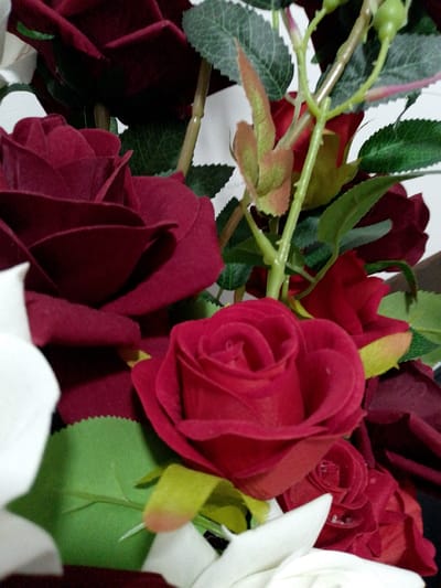 The image shows a close-up of a bouquet of artificial red roses and green leaves against a white background. The roses are of varying shades of red, with some appearing deep crimson and others a brighter red. The petals are intricately detailed, suggesting a realistic artificial flower. The leaves are a vibrant green, with a textured surface. The bouquet is tightly arranged, with several roses overlapping and some buds visible. There are no people, actions, or interactions depicted in the image. The environment appears to be indoors, with a plain white background that suggests a studio setting or a room with neutral decor. The location in the city of Shirin, Uzbekistan, is not visually represented in the image; there are no identifying landmarks, architecture, or geographical features that would indicate this specific location.