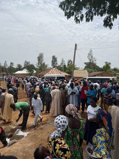 A large crowd has gathered outside in Sabon Gida, Nigeria. Most people are standing and facing in the same direction, many are wearing traditional African clothing. A white bus is parked along the edge of the gathering, near several buildings with metal roofs. An electrical pole with lines is visible. The ground is dirt. Several people are digging into a large white bag containing a dark substance, possibly soil or fertilizer. The sky is overcast.