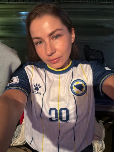 A young woman with blonde hair and blue eyes takes a selfie. She is wearing a white and blue soccer jersey with yellow trim and the number "20" in blue on the front. The jersey also features a crest with the outline of Bosnia and Herzegovina. The woman is outdoors at night, with a blurry background that suggests a city street. There are no explicit indications of the city of Travnik, Bosnia and Herzegovina within the image itself.
